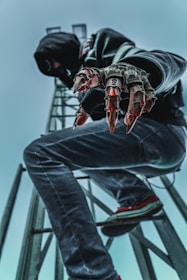A person dressed in dark clothing, including a hood, is crouching on a metal structure. They have mechanical, claw-like gloves extending from their hands, giving a futuristic or cybernetic appearance. The viewpoint is from below, with an emphasis on the person's gloves and legs, set against a cloudy sky.