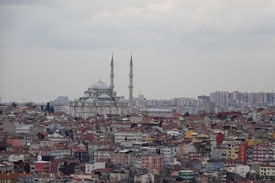 A panoramic view of a city with a large mosque featuring two tall minarets, surrounded by numerous tightly-packed residential and commercial buildings. The sky is overcast with grey clouds, creating a subdued light over the cityscape. In the background, there are additional high-rise buildings and urban structures.