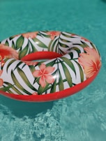 A colorful inflatable ring with a floral pattern floats on the clear blue water of a pool. The ring is decorated with orange and pink flowers and green leaves, creating a vibrant contrast against the water.