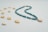 A necklace composed of blue beads and gold accents is gracefully arranged in a curving shape. Scattered around the necklace are several small, beige cowrie shells on a light, neutral background.