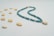 A necklace composed of blue beads and gold accents is gracefully arranged in a curving shape. Scattered around the necklace are several small, beige cowrie shells on a light, neutral background.