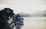 Several rustic houses sit on the edge of a tranquil lake, surrounded by lush trees. Misty mountains form the backdrop under a cloudy sky, creating a serene and peaceful atmosphere.