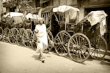 An elderly person walks along a street lined with vintage rickshaws. The rickshaws have large wooden wheels and are covered with faded canopies. The setting appears to be a city street with a series of old buildings and signs in the background.