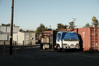 A white truck with a blue tarp covering the cab is parked next to large shipping containers in an industrial area. Several yellow poles are standing nearby, and a wooden utility pole is present. In the background, there are trees and distant hills under a clear blue sky.