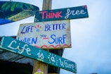 Several colorful wooden signs attached to a pole with motivational and surfing-related messages. The messages read, 'Think free,' 'Life is better when you surf,' and 'Life is a wave.' The signs are painted in various shades of blue, green, pink, and white against a clear sky.