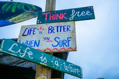 Several colorful wooden signs attached to a pole with motivational and surfing-related messages. The messages read, 'Think free,' 'Life is better when you surf,' and 'Life is a wave.' The signs are painted in various shades of blue, green, pink, and white against a clear sky.