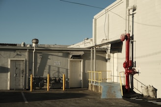An industrial area features a building with a 'Reserved Parking' sign on the wall. There is a bright red pipe system attached to the structure and yellow safety poles are positioned in front. A small ramp with a blue surface and security bars around it is also visible. The scene is bathed in warm, late-afternoon sunlight, creating distinct shadows.