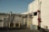 An industrial area features a building with a 'Reserved Parking' sign on the wall. There is a bright red pipe system attached to the structure and yellow safety poles are positioned in front. A small ramp with a blue surface and security bars around it is also visible. The scene is bathed in warm, late-afternoon sunlight, creating distinct shadows.