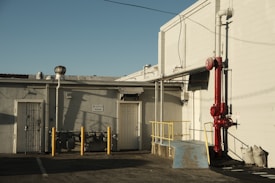 An industrial area features a building with a 'Reserved Parking' sign on the wall. There is a bright red pipe system attached to the structure and yellow safety poles are positioned in front. A small ramp with a blue surface and security bars around it is also visible. The scene is bathed in warm, late-afternoon sunlight, creating distinct shadows.