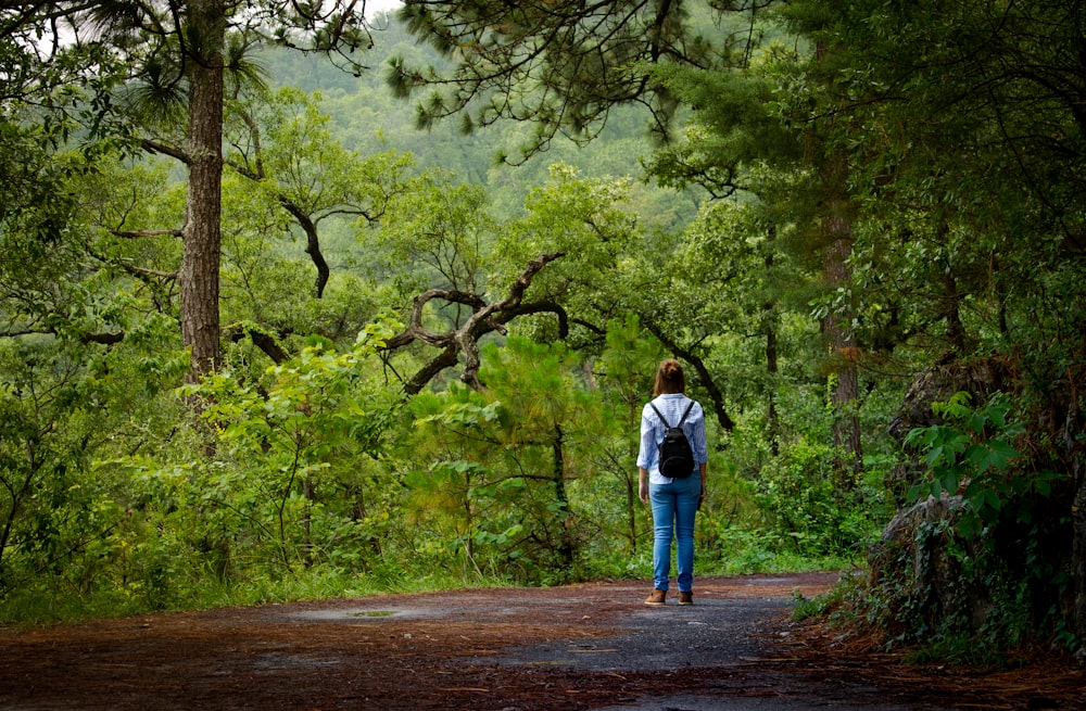 mulher em jaqueta jeans azul e jeans azul andando no caminho entre árvores verdes durante