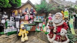 A whimsical display featuring a variety of plush toys, figurines, and decorations. The scene includes a large festive Santa Claus figure in red holding presents, surrounded by numerous stuffed animals like sheep and dogs. Colorful cartoon characters and other playful decorations adorn a rustic wooden stall, enhanced by greenery and festive motifs.