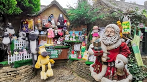 A whimsical display featuring a variety of plush toys, figurines, and decorations. The scene includes a large festive Santa Claus figure in red holding presents, surrounded by numerous stuffed animals like sheep and dogs. Colorful cartoon characters and other playful decorations adorn a rustic wooden stall, enhanced by greenery and festive motifs.