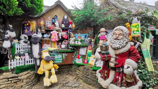 A whimsical display featuring a variety of plush toys, figurines, and decorations. The scene includes a large festive Santa Claus figure in red holding presents, surrounded by numerous stuffed animals like sheep and dogs. Colorful cartoon characters and other playful decorations adorn a rustic wooden stall, enhanced by greenery and festive motifs.