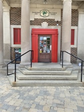 A brick building entrance with a bold red-framed door is flanked by two large pillars. The entrance has a set of stone steps leading up to it with black metal railings on either side. A sign above the door indicates the establishment's name and location. The design includes red window shutters to the side.