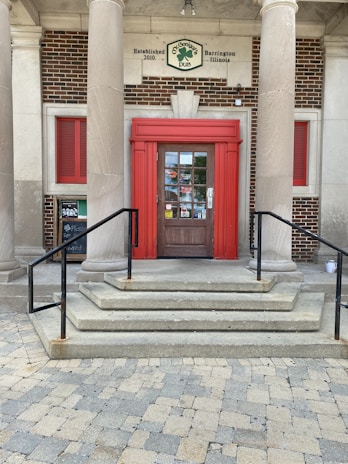 A brick building entrance with a bold red-framed door is flanked by two large pillars. The entrance has a set of stone steps leading up to it with black metal railings on either side. A sign above the door indicates the establishment's name and location. The design includes red window shutters to the side.