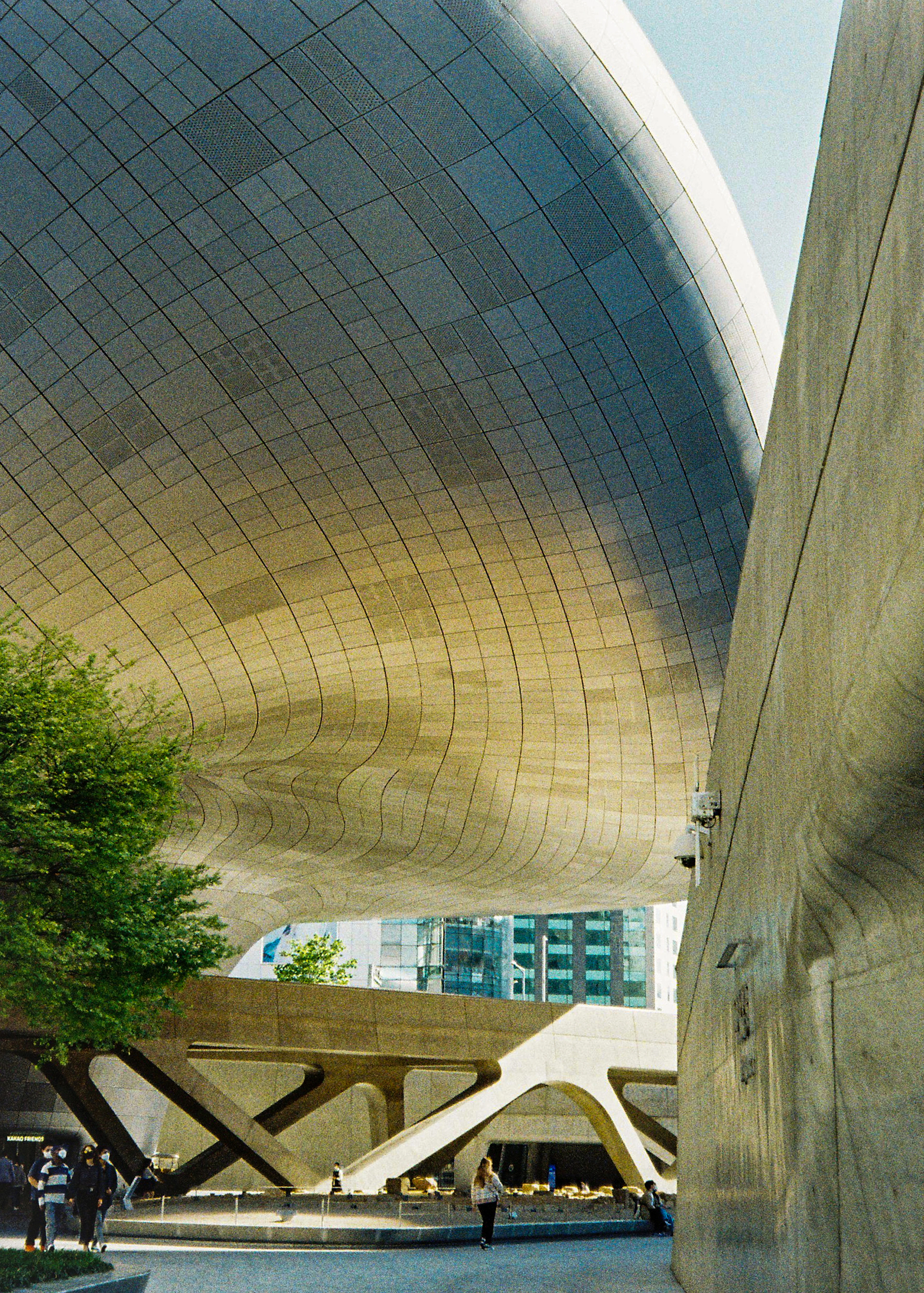 The curving silver aluminium underside of the Dongdaemun Design Plaza in Seoul, with people crossing the plaza beneath it