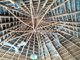 The image displays a wooden ceiling with a geometric pattern, where multiple beams radiate from a central point. The ceiling is equipped with two ceiling fans and several light fixtures that hang down, dispersed around the space. The background includes dark insulation material between the wooden beams.
