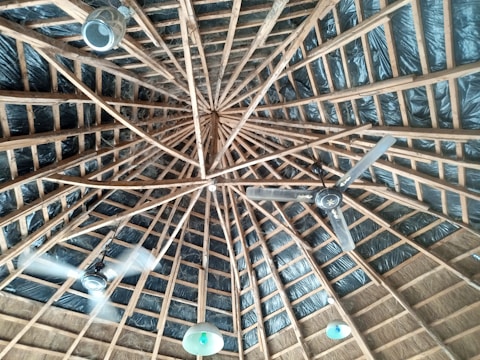 The image displays a wooden ceiling with a geometric pattern, where multiple beams radiate from a central point. The ceiling is equipped with two ceiling fans and several light fixtures that hang down, dispersed around the space. The background includes dark insulation material between the wooden beams.
