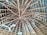 The image displays a wooden ceiling with a geometric pattern, where multiple beams radiate from a central point. The ceiling is equipped with two ceiling fans and several light fixtures that hang down, dispersed around the space. The background includes dark insulation material between the wooden beams.