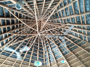 The image displays a wooden ceiling with a geometric pattern, where multiple beams radiate from a central point. The ceiling is equipped with two ceiling fans and several light fixtures that hang down, dispersed around the space. The background includes dark insulation material between the wooden beams.