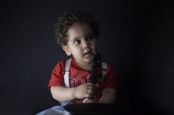 A child with curly hair is sitting, holding an object resembling a stick or microphone. The child is wearing a red shirt with white suspenders, and the background is dark, providing contrast to the child in the foreground.