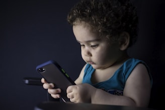A young child with curly hair is intently looking at a smartphone they are holding. The child is wearing a blue sleeveless shirt and is seated against a dark background, creating a soft contrast. The scene is dimly lit, highlighting the child's focused expression.