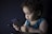 A young child with curly hair is intently looking at a smartphone they are holding. The child is wearing a blue sleeveless shirt and is seated against a dark background, creating a soft contrast. The scene is dimly lit, highlighting the child's focused expression.