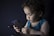 A young child with curly hair is intently looking at a smartphone they are holding. The child is wearing a blue sleeveless shirt and is seated against a dark background, creating a soft contrast. The scene is dimly lit, highlighting the child's focused expression.