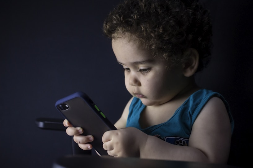 A young child with curly hair is intently looking at a smartphone they are holding. The child is wearing a blue sleeveless shirt and is seated against a dark background, creating a soft contrast. The scene is dimly lit, highlighting the child's focused expression.