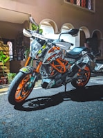 A sports motorcycle with vibrant orange and white accents is parked on an asphalt surface in front of a building. The bike has multiple stickers and detailed graphics. There are plants and decorations visible in the background near an entrance with arched doorways.