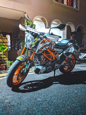 A sports motorcycle with vibrant orange and white accents is parked on an asphalt surface in front of a building. The bike has multiple stickers and detailed graphics. There are plants and decorations visible in the background near an entrance with arched doorways.