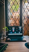 A computer screen showcases a virtual meeting with several participants displayed on it. The laptop is on a wooden desk near a window with a crisscross pattern and an outdoor scene visible through it, including plants and buildings. There are small plants and a bottle with water on the desk beside the laptop.