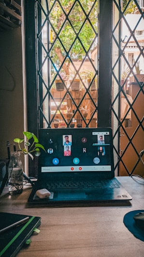 A computer screen showcases a virtual meeting with several participants displayed on it. The laptop is on a wooden desk near a window with a crisscross pattern and an outdoor scene visible through it, including plants and buildings. There are small plants and a bottle with water on the desk beside the laptop.
