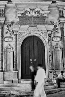 An ornate architectural doorway featuring intricate carvings and designs, framed by large stone columns. Arabic script adorns the top panel, highlighting its historical or cultural significance. Two blurred figures in motion pass by in the foreground. On the right side of the steps, two seated individuals appear to be in conversation.