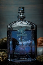 A clear glass perfume bottle, partially covered with water droplets, stands amidst smooth, multi-colored stones. The bottle contains a blue liquid and has detailed engravings and a spray nozzle at the top.