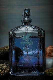 A clear glass perfume bottle, partially covered with water droplets, stands amidst smooth, multi-colored stones. The bottle contains a blue liquid and has detailed engravings and a spray nozzle at the top.