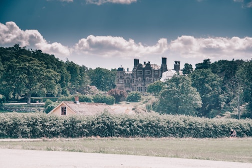 A grand, historic mansion with numerous chimneys and intricate architecture sits atop a hill surrounded by dense greenery. In the foreground, a smaller, rustic house with a thatched roof is nestled amidst hedges and trees. The sky is partly cloudy, casting a serene yet slightly moody atmosphere over the landscape.