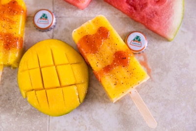 A vibrant, tropical scene with a sliced mango showing its juicy yellow flesh and a mango-flavored popsicle drizzled with chili powder and sauce. Two bottle caps from a Jarritos beverage are placed near the fruit, and a wedge of watermelon adds a splash of red to the composition.