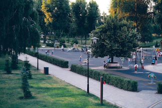A park scene with children and adults enjoying various outdoor activities. There is a large playground area with play structures and equipment. People are scattered throughout the park, engaged in walking, playing, and sitting on benches. Lush greenery surrounds the area with trees and trimmed bushes lining the pathways. The scene suggests a typical day at a community park.