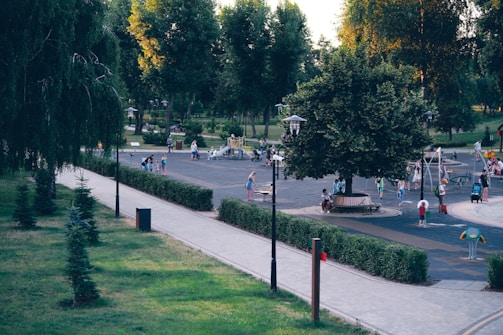 A park scene with children and adults enjoying various outdoor activities. There is a large playground area with play structures and equipment. People are scattered throughout the park, engaged in walking, playing, and sitting on benches. Lush greenery surrounds the area with trees and trimmed bushes lining the pathways. The scene suggests a typical day at a community park.