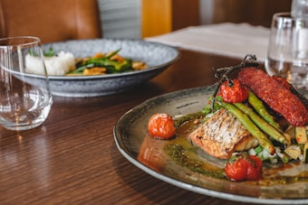 A well-presented dish featuring grilled fish, asparagus, roasted cherry tomatoes, and a slice of red sausage on a ceramic plate with a green sauce. Another plate with rice and vegetables is slightly blurred in the background. The table setting includes clear glassware and is arranged on a wooden table.