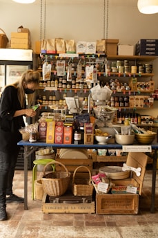 A person wearing dark clothing and a mask examines a green item in a small, cozy store. The store features a wooden table covered with various products, including packaged food items in colorful boxes, jars, and baskets. Shelves filled with more products line the wall behind the table, displaying jars, boxes, and bottles. Two hanging lights illuminate the area, giving it a warm and inviting feel.