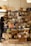 A person wearing dark clothing and a mask examines a green item in a small, cozy store. The store features a wooden table covered with various products, including packaged food items in colorful boxes, jars, and baskets. Shelves filled with more products line the wall behind the table, displaying jars, boxes, and bottles. Two hanging lights illuminate the area, giving it a warm and inviting feel.