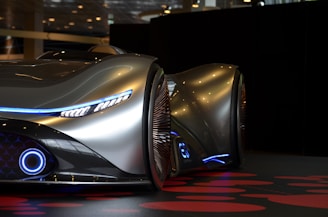 A sleek, futuristic car with a metallic silver finish and smooth, aerodynamic curves. It has distinctive blue LED lighting accents, including strip lights along the front and around the wheels. The car features advanced design elements and appears to be showcased in an indoor environment with a dimly lit, modern backdrop.
