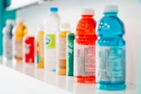 A row of assorted beverage bottles is displayed on a white shelf. The bottles vary in color and type, featuring bright shades of red, blue, yellow, and green among others. They include plastic sports drinks and bottled water, some with visible nutrition labels.