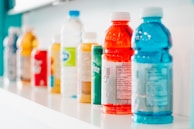 A row of assorted beverage bottles is displayed on a white shelf. The bottles vary in color and type, featuring bright shades of red, blue, yellow, and green among others. They include plastic sports drinks and bottled water, some with visible nutrition labels.
