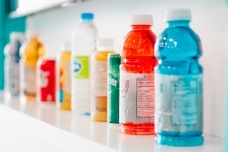 A row of assorted beverage bottles is displayed on a white shelf. The bottles vary in color and type, featuring bright shades of red, blue, yellow, and green among others. They include plastic sports drinks and bottled water, some with visible nutrition labels.