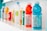 A row of assorted beverage bottles is displayed on a white shelf. The bottles vary in color and type, featuring bright shades of red, blue, yellow, and green among others. They include plastic sports drinks and bottled water, some with visible nutrition labels.
