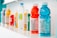 A row of assorted beverage bottles is displayed on a white shelf. The bottles vary in color and type, featuring bright shades of red, blue, yellow, and green among others. They include plastic sports drinks and bottled water, some with visible nutrition labels.