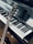 A close-up view of a piano keyboard with a guitar resting against it. The piano keys are clean and polished, reflecting light. The guitar, positioned diagonally, has a wooden body and the brand name is visible on the headstock.